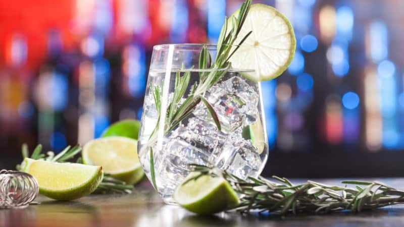 Nahaufnahme von einem Glas mit Gin und Gurken in einer Bar. Daneben liegen Limetten und Kräuter.