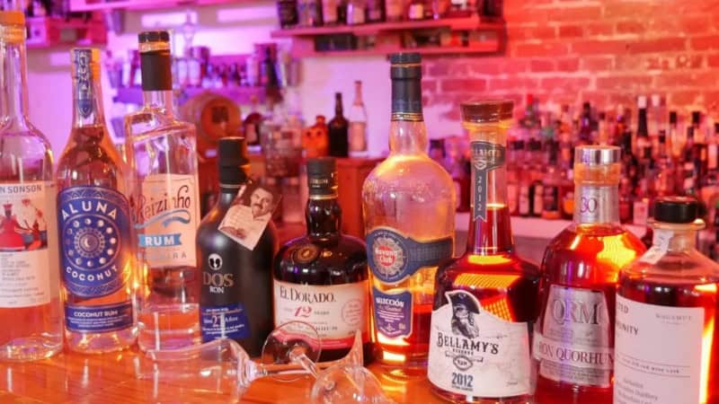 Rum-Flaschen stehen aufgereiht auf einem farbig beleuchteten Bartresen. Davor liegen zwei Gläser.