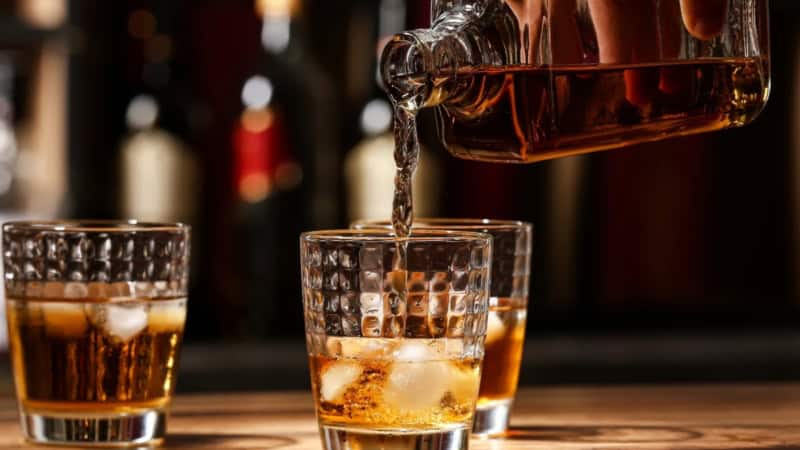 Rum wird aus einer Flasche in ein Glas gefüllt. Zwei volle Gläser mit Rum und Eiswürfeln stehen daneben.