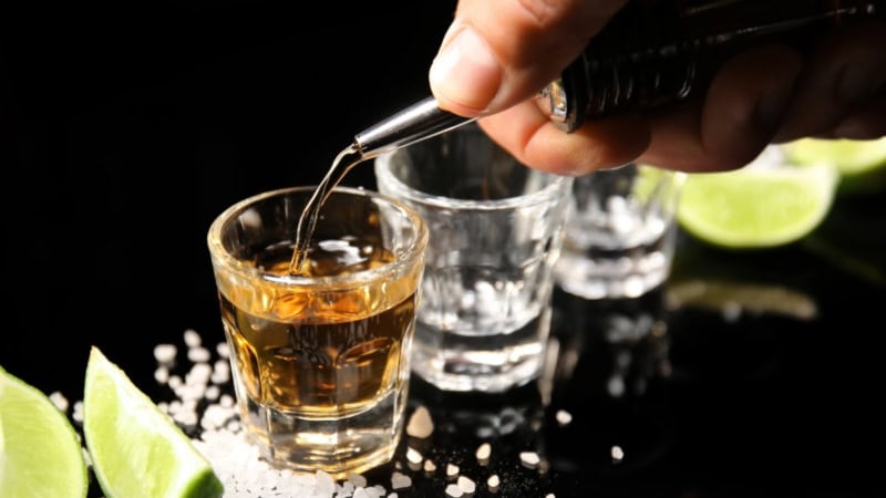 Drei Shotgläser, Salz und Limetten liegen auf einem dunklen Untergrund. In ein Glas wird Tequila eingefüllt.