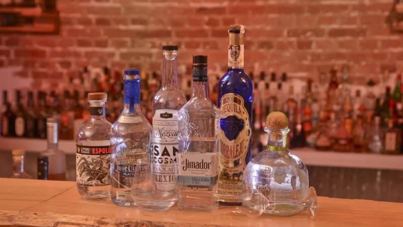 Tequila-Flaschen und ein Glas stehen aufgereiht auf einem bunt beleuchteten Bartresen.