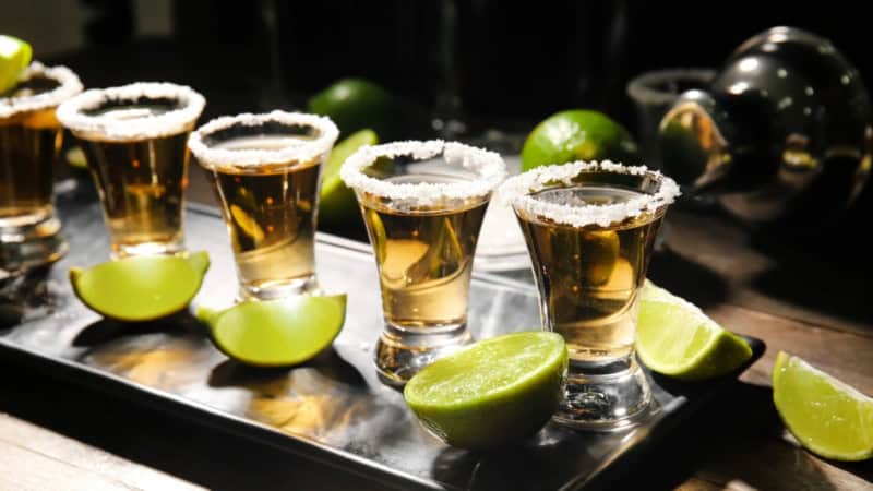 Tequila Shots stehen aufgereiht auf einer Unterlage. Daneben liegen Limetten.