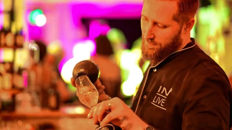 Barkeeper steht hinter einer Bar und schüttet Champagner in ein Sektglas.