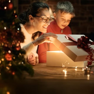 Eine Frau und ihr Kind knien vor dem Weihnachtsbaum und heben den Deckel einer Geschenkbox an.