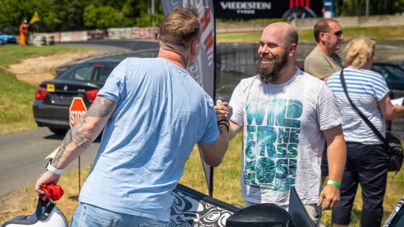 Zwei Männer stehen vor einem Auto an einer Rennstrecke und begrüßen sich.