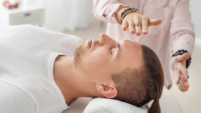 Ein Mann liegt auf einer Liege und eine Therapeutin hält ihre Hände über seinen Kopf und gibt ihm Energie.