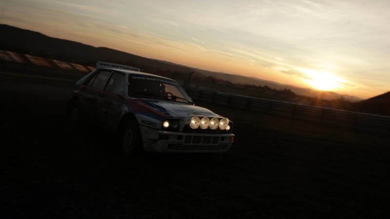 Ein weißes Sportauto mit roten und blauen Details driftet bei Sonnenuntergang in einer Kurve auf dem Nürburgring.