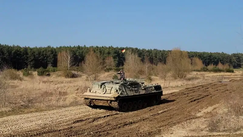 Ein Mann der mit einem Panzer über einen Matsch Weg fährt.