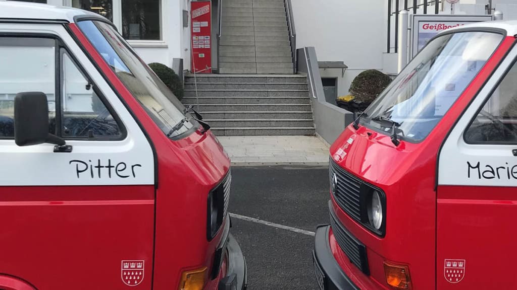 VW-Bulli-Tour-2-bullies-gegenüber 2 rote VW Bullies stehen sich frontal gegenüber.