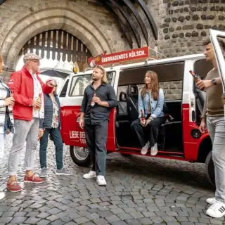 Ein VW Bulli der vor einem Burgen Tor in Köln steht. Die tour Teilnehmer stehen vor dem Bulli und hörem dem Guide zu während sie ein Bier trinken.