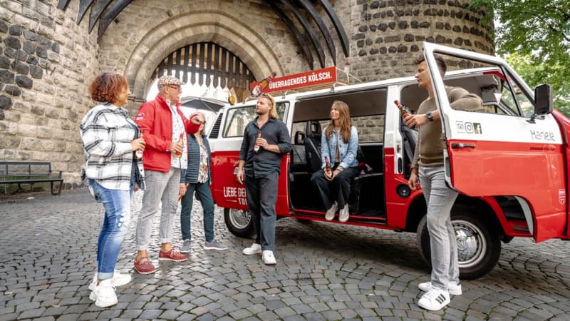 Ein VW Bulli der vor einem Burgen Tor in Köln steht. Die tour Teilnehmer stehen vor dem Bulli und hörem dem Guide zu während sie ein Bier trinken.