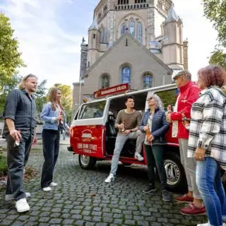 Ein VW Bulli der vor einer Kirche parkt. Der Tourguide erzählt den VW Bulli Tourteilnehmern etwas während diese ein Bier trinken.