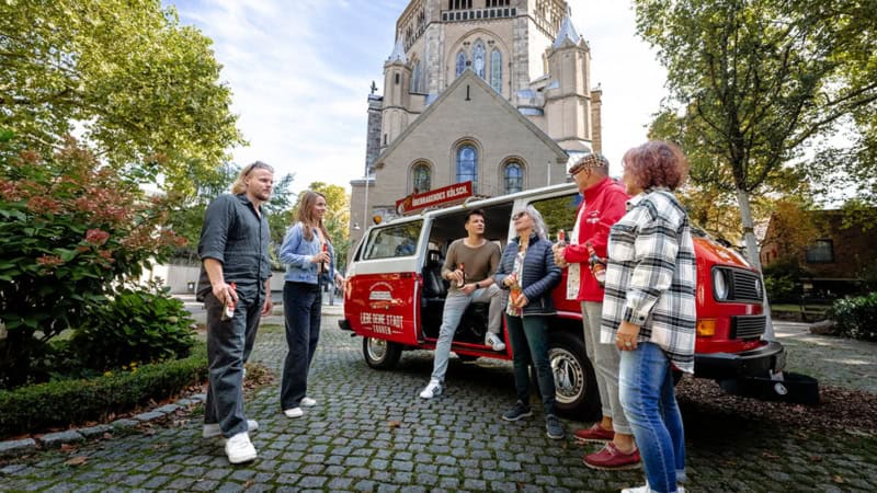 Ein VW Bulli der vor einer Kirche parkt. Der Tourguide erzählt den VW Bulli Tourteilnehmern etwas während diese ein Bier trinken.