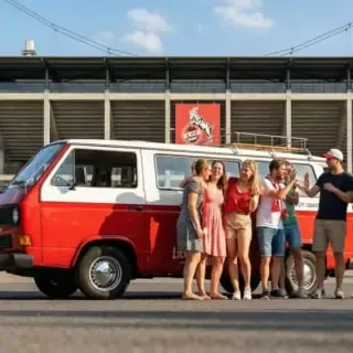 Eine gruppe VW Bulli Tour Teilnehmer steht mit dem Bulli vor dem FC Kölln Stadion und freut sich.