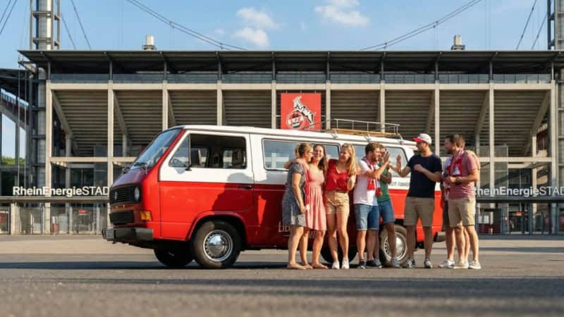 Eine gruppe VW Bulli Tour Teilnehmer steht mit dem Bulli vor dem FC Kölln Stadion und freut sich.