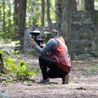 Person hockt mit Paintballgewehr in der Hand im Wald und zielt auf einen Gegenspieler