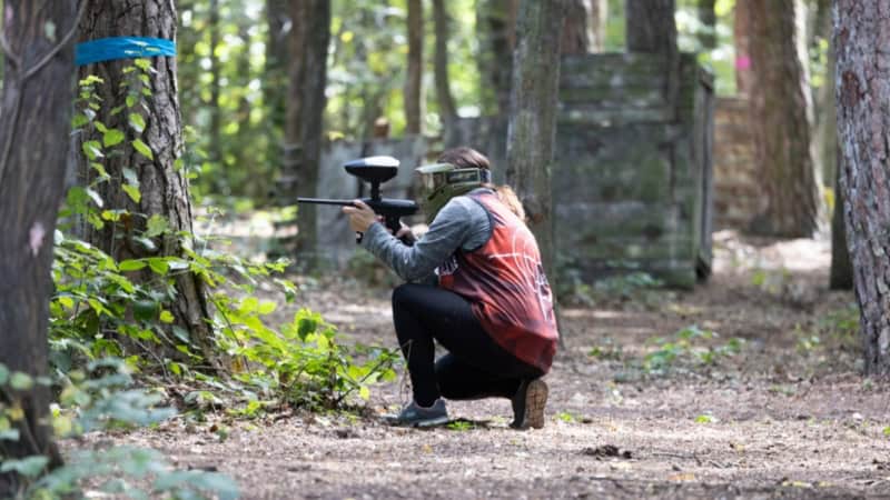 Person hockt mit Paintballgewehr in der Hand im Wald und zielt auf einen Gegenspieler