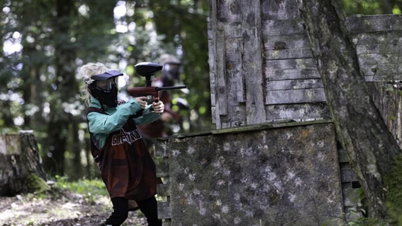 Paintballspieler mit Schutzmaske zielt mit einem Markierer an einem Baum vorbei