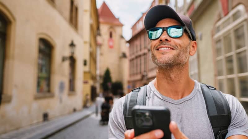 Lächelnder Mann mit Sonnenbrille und Rucksack navigiert per Smartphone durch die historische Altstadtgasse - digitale Stadtrallye.