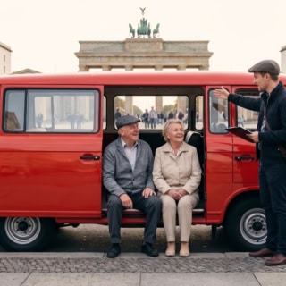 Ein älteres Paar sitzt in einem roten Oldtimer Barkas vor dem Brandenburger Tor in Berlin - DDR Stadtrundfahrt.